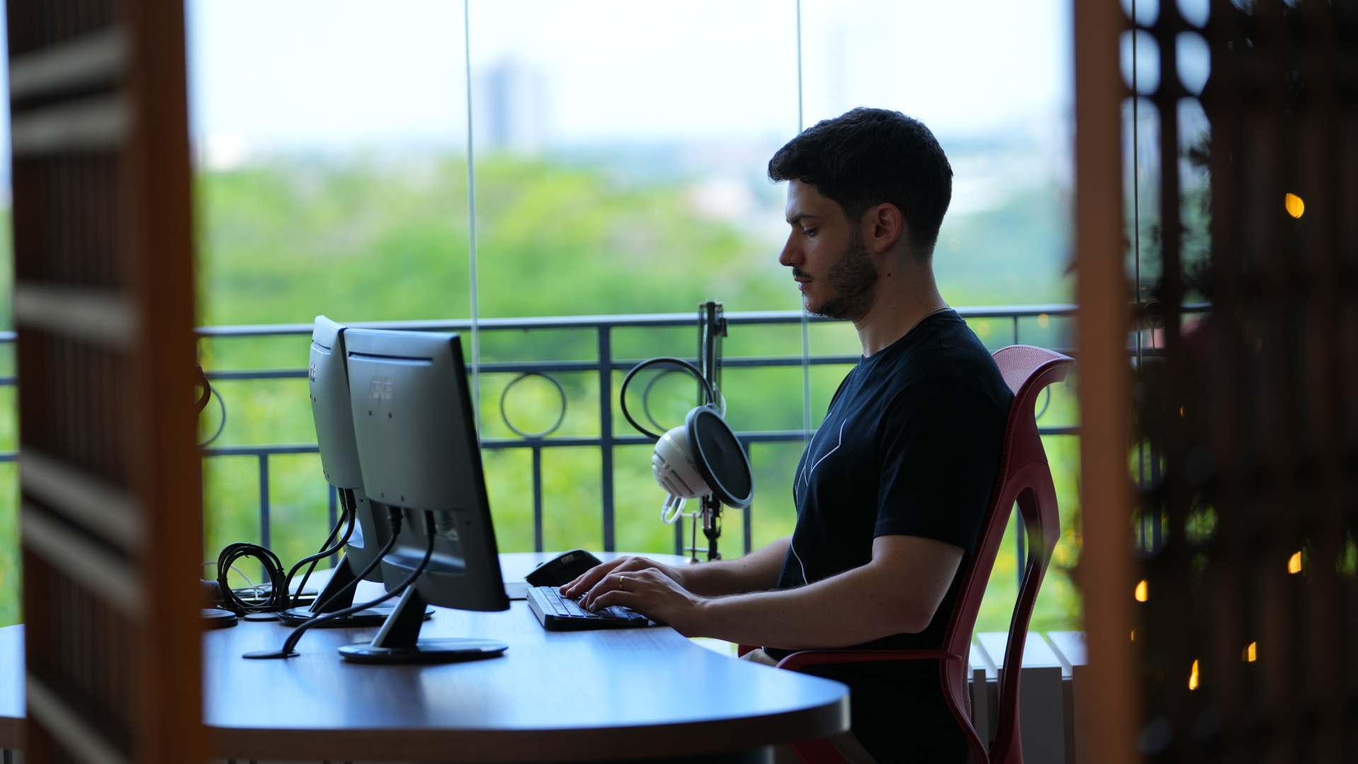 Andre is sitting at a desk with dual monitors. A microphone is set up beside the desk, with a green and serene outdoor background visible through large windows